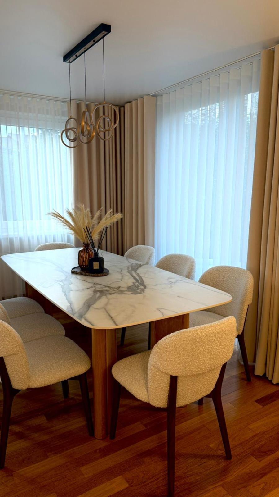 Dining room with marble table and beige chairs, large windows with curtains.