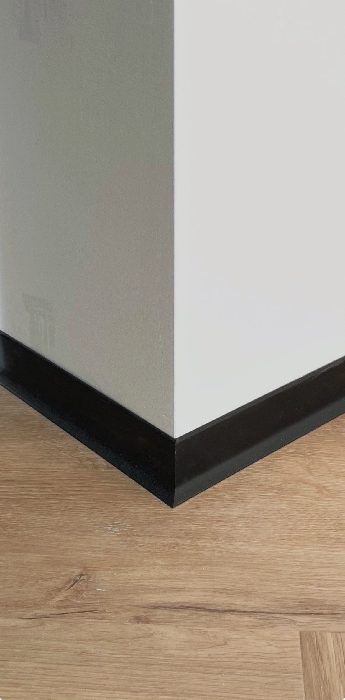 Close-up of a corner where a white wall meets a wooden floor with black aluminium baseboard.